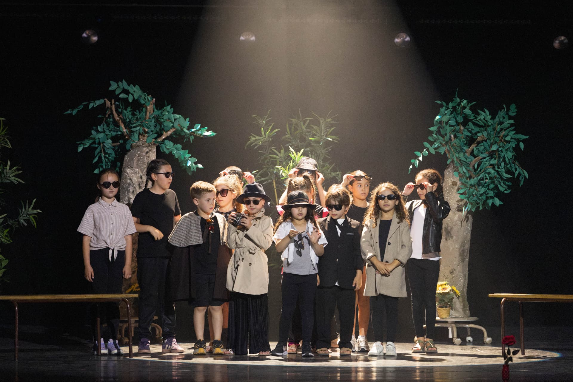 Un groupe d'enfants du Labo Théâtre en scène, lunettes de soleil et chapeaux, sous un projecteur